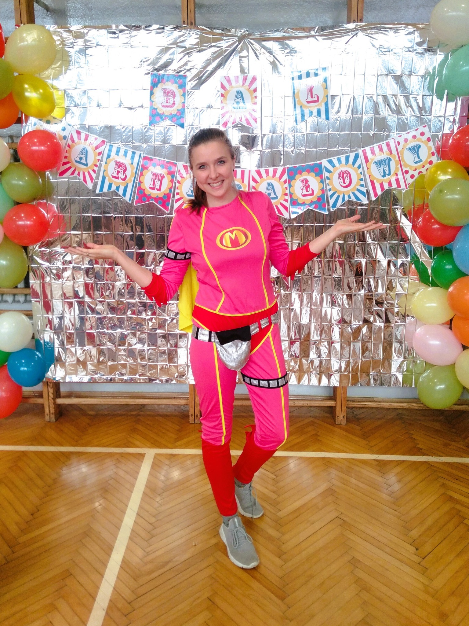 Animator w różowym kostiumie superbohatera z literą 'M' na tle srebrnej ściany z balonami i napisem 'Happy Birthday' w Orzeszu, z rozłożonymi rękami i uśmiechem.