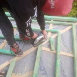 Pracownik w czarnej bluzie z kapturem i sportowych butach przycina deskę piłą elektryczną na konstrukcji dachu pokrytej membraną.