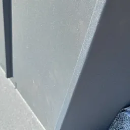 Szare, metalowe obróbki blacharskie na dachu, widoczne buty robocze i zwinięty materiał izolacyjny w tle.