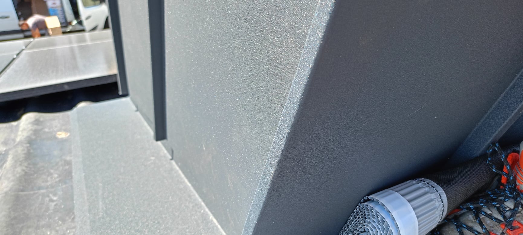 Szare, metalowe obróbki blacharskie na dachu, widoczne buty robocze i zwinięty materiał izolacyjny w tle.