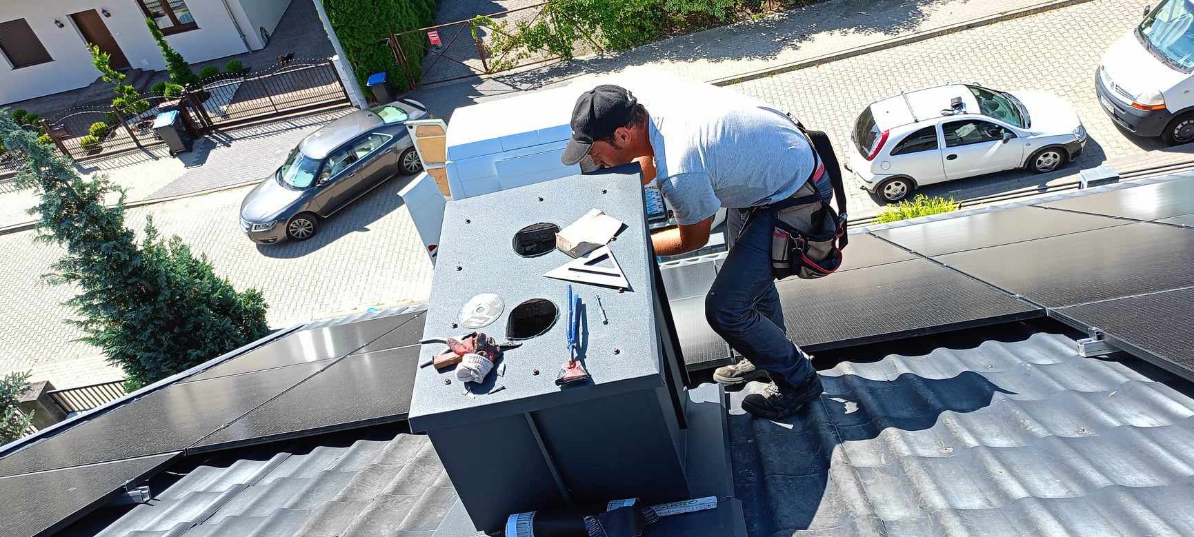Pracownik w trakcie montażu kominka wentylacyjnego na dachu pokrytym panelami fotowoltaicznymi i blachodachówką, widoczne narzędzia pracy i samochody w tle.