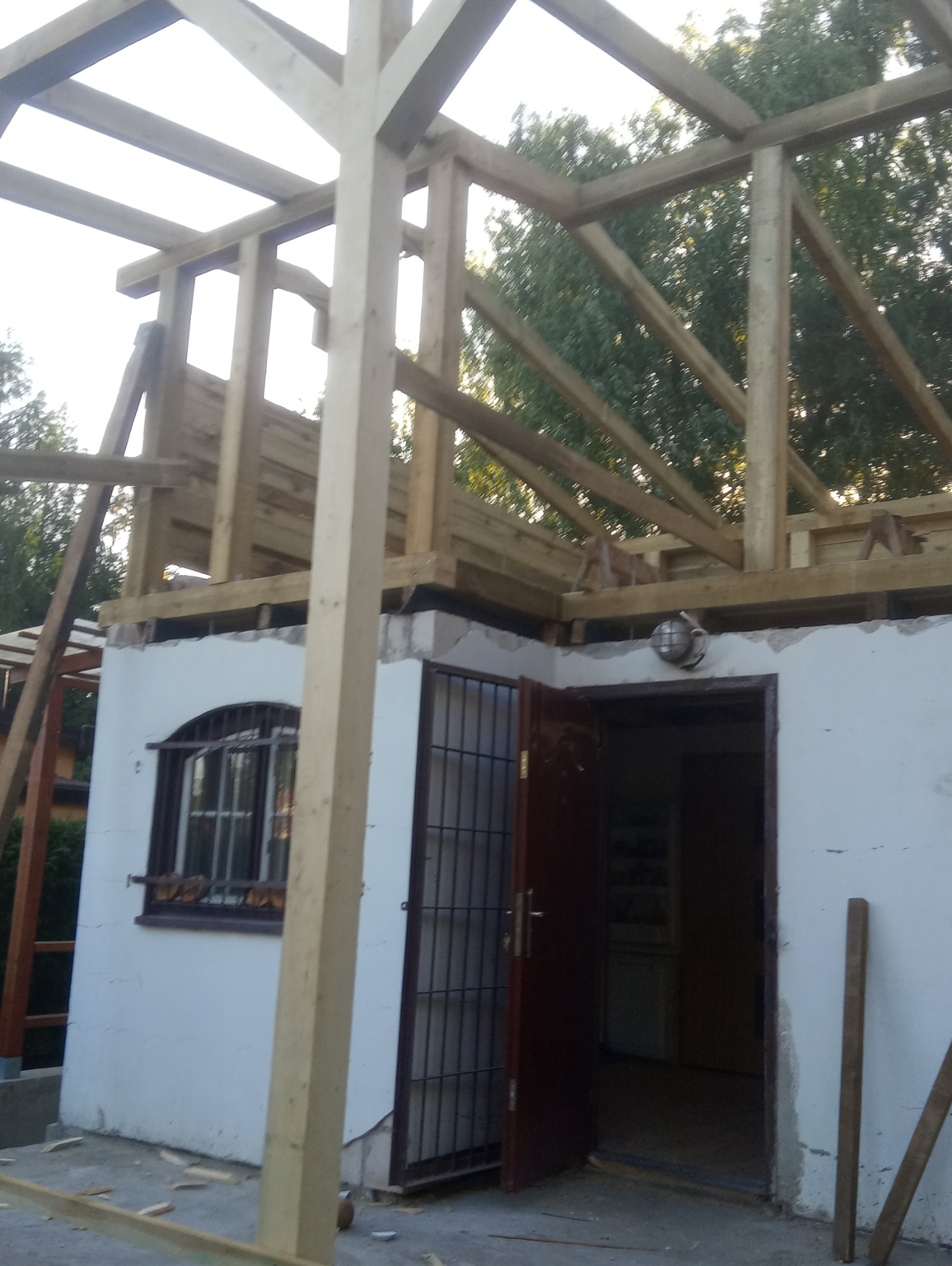 Konstrukcja drewniana nowego dachu w trakcie budowy, widok na biały budynek z oknem i otwartymi drzwiami, drzewa w tle.