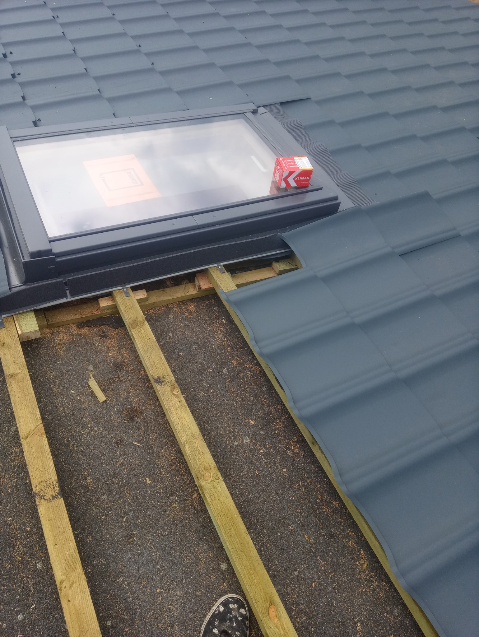 Montaż okna dachowego w trakcie krycia dachu blachodachówką w kolorze grafitowym, widoczne drewniane łaty i pudełko z akcesoriami montażowymi.