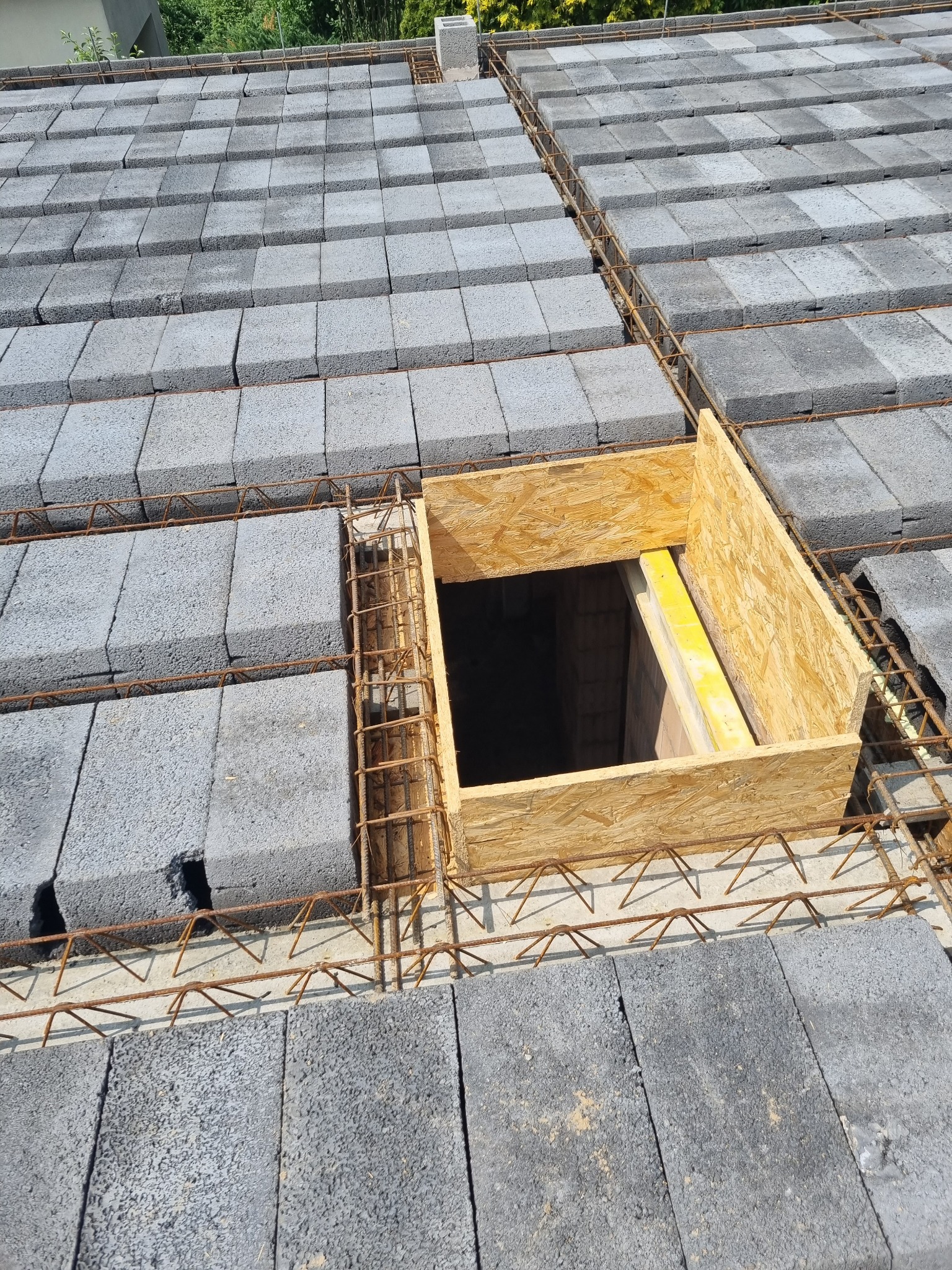 Wiązanie zbrojenia na stropie z bloczków betonowych, widoczny otwór szalunkowy z desek, w tle zieleń drzew.