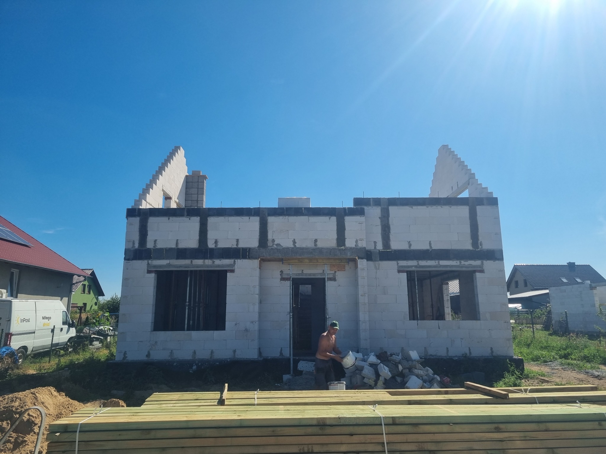 Wznoszenie ścian budynku z betonu komórkowego; widoczny murarz w trakcie pracy, paleta z drewnem na pierwszym planie, błękitne niebo w tle.