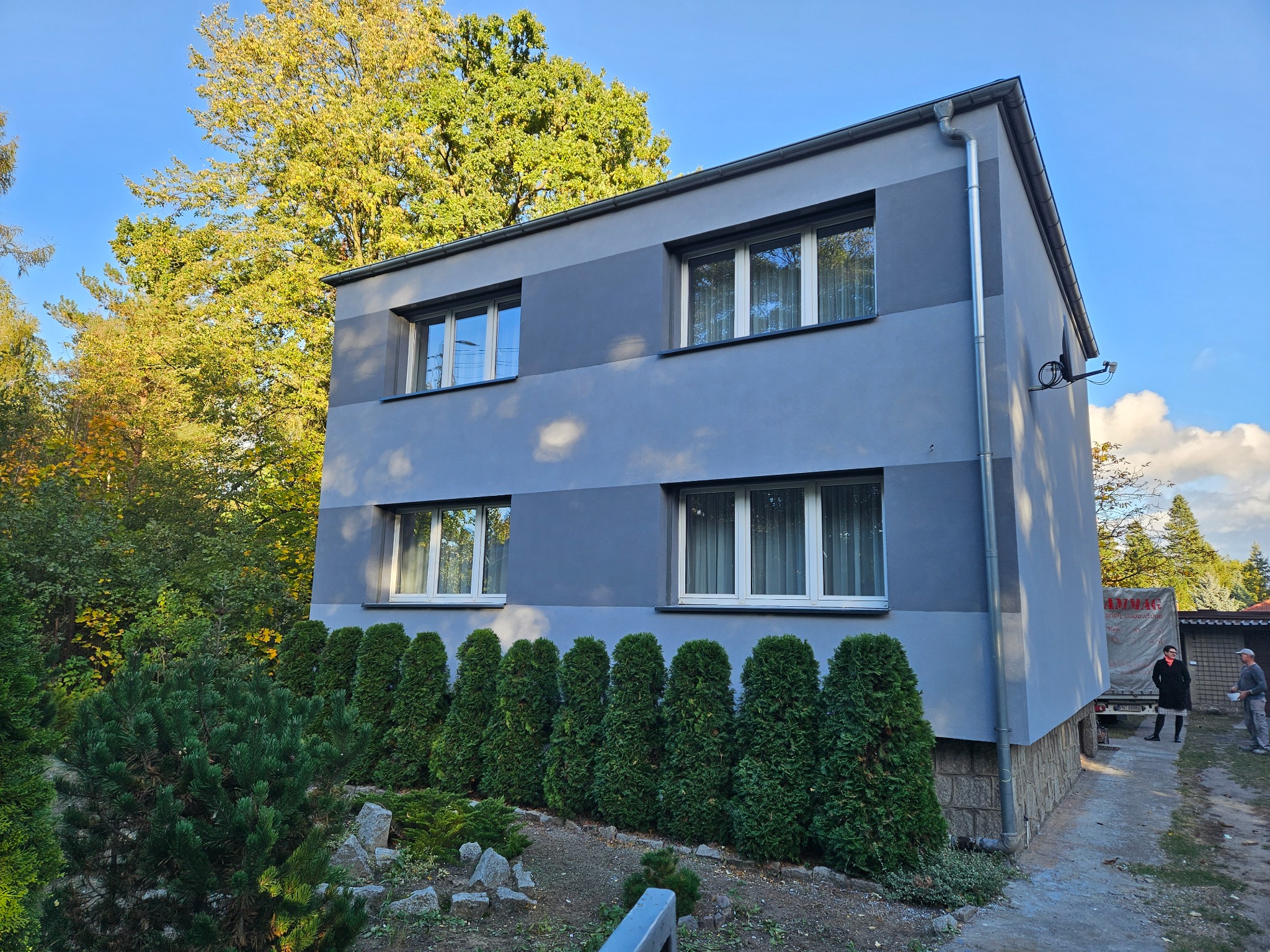 Dwukondygnacyjny dom murowany z szarą elewacją, widoczne okna z białymi ramami, rząd tujek przed domem, fragment elewacji wykończony kamieniem, na bocznej ścianie antena satelitarna