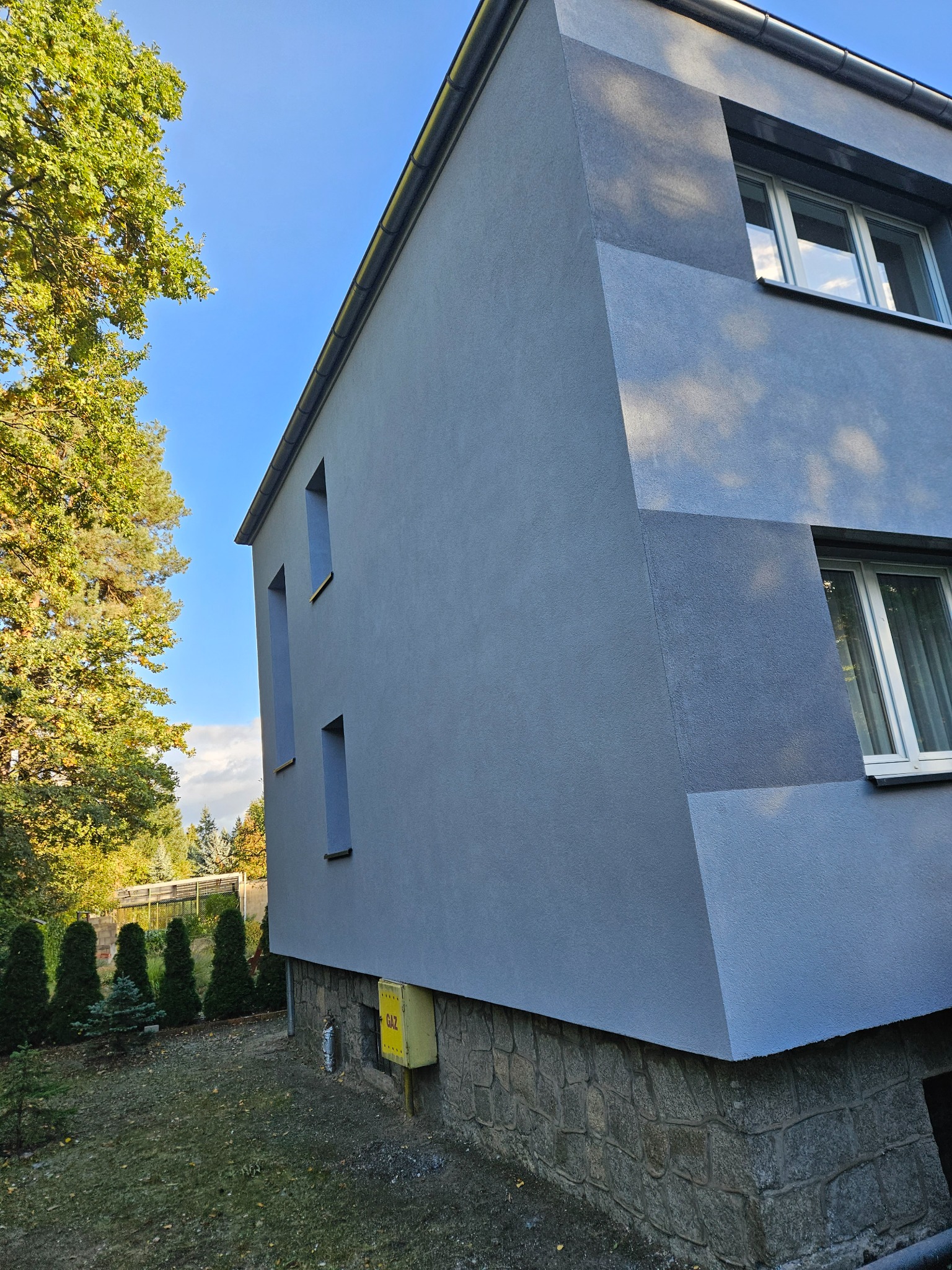 Dwukondygnacyjny dom z szarą elewacją w odcieniach, z widocznymi oknami i podmurówką z kamienia, otoczony zielenią i drzewami.