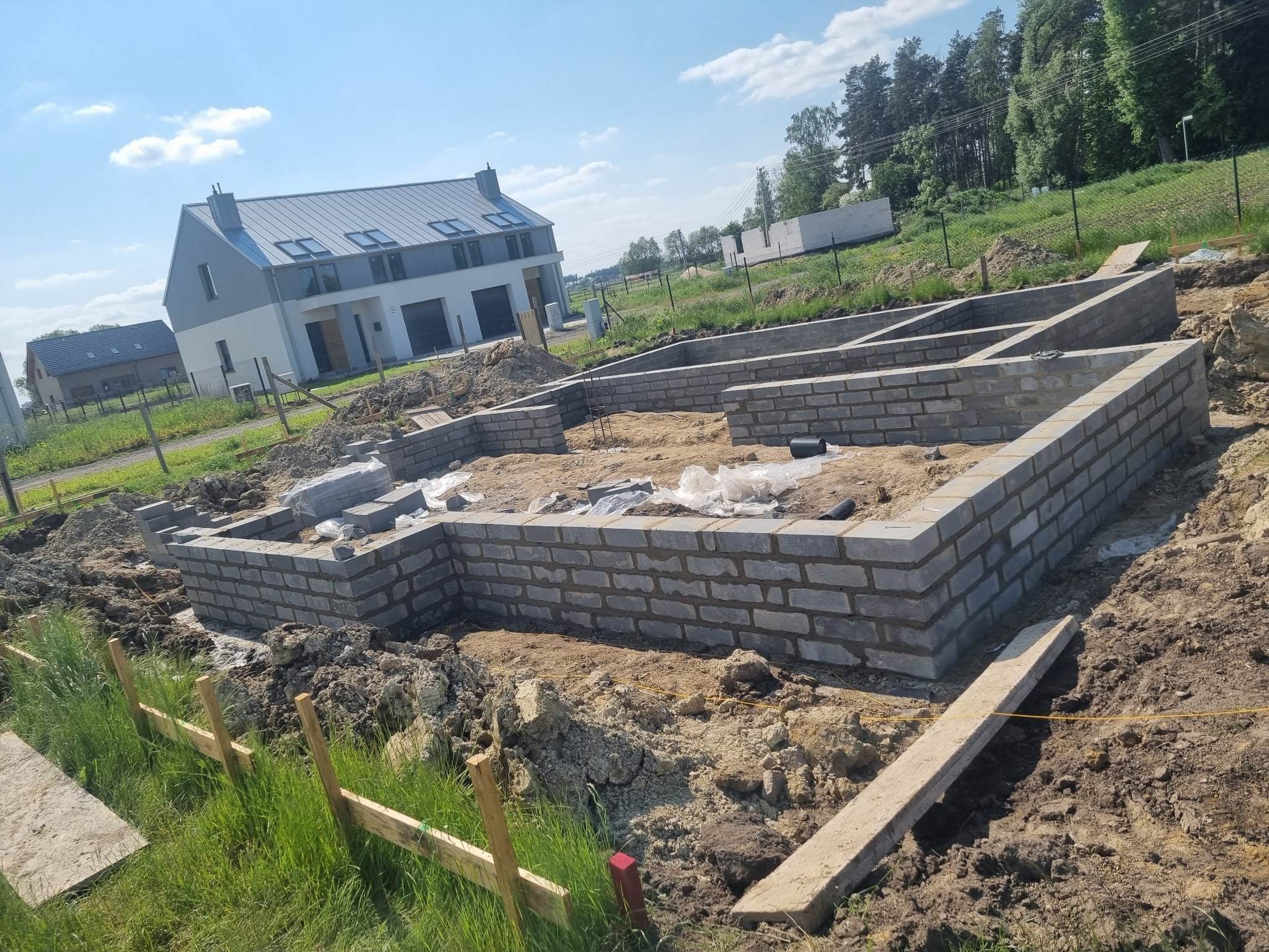 Wznoszenie fundamentów z bloczków betonowych na terenie budowy domu jednorodzinnego, widoczne zbrojenie i warstwy izolacyjne, w tle nowoczesny dom z szarą blachodachówką.