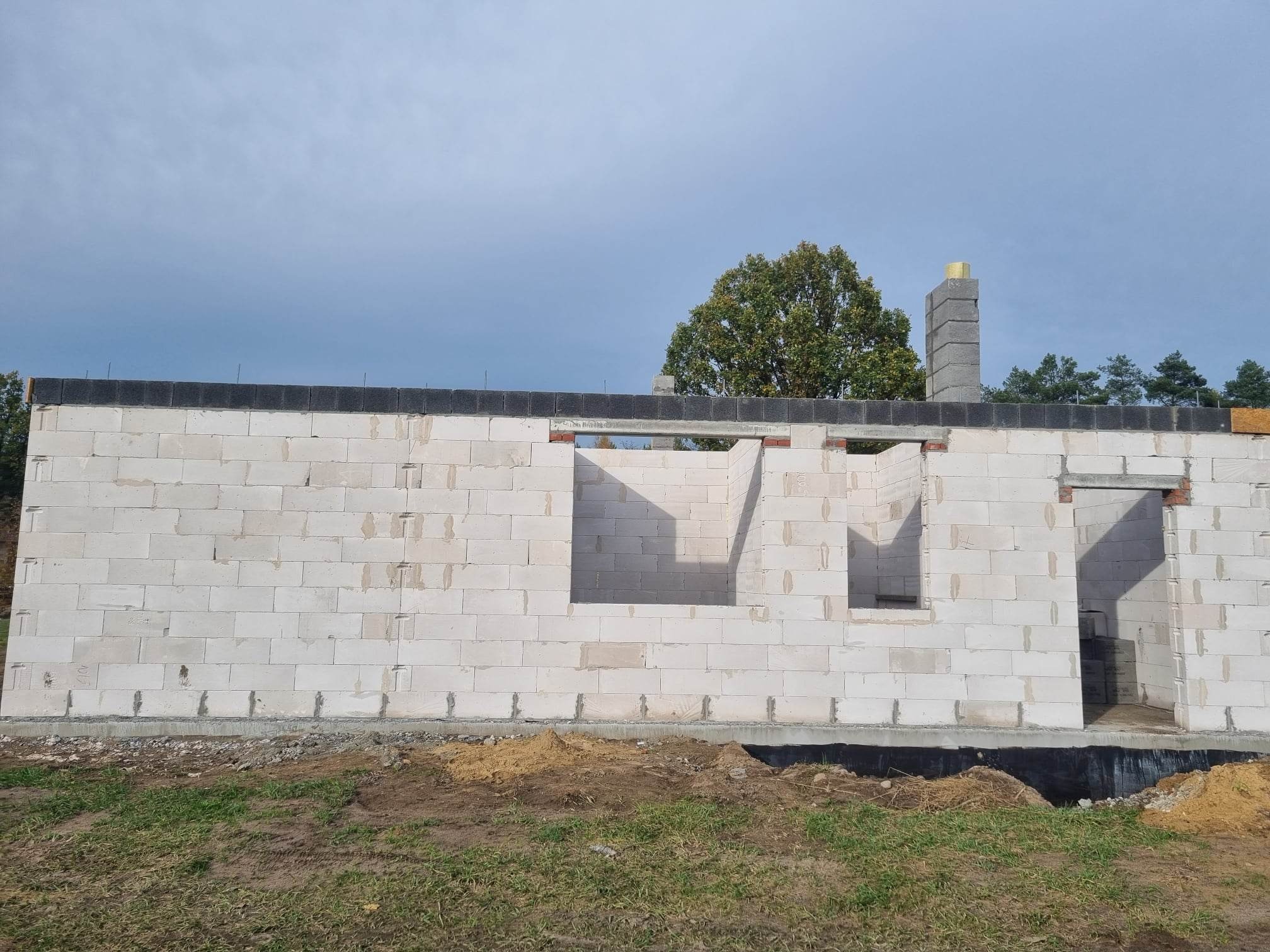 Wznoszone ściany budynku z betonu komórkowego, widoczne otwory okienne i drzwiowe, fragment komina z szarej cegły, teren wokół budynku z fragmentami trawy i ziemi.