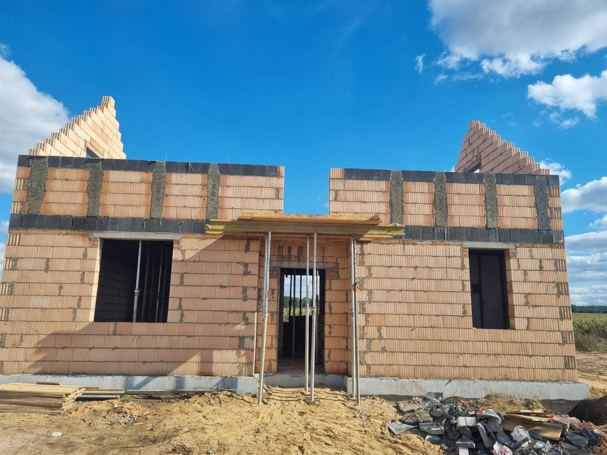 Wznoszona ściana zewnętrzna domu z pustaków ceramicznych, z widocznymi otworami okiennymi i zbrojonym wieńcem, na tle błękitnego nieba.