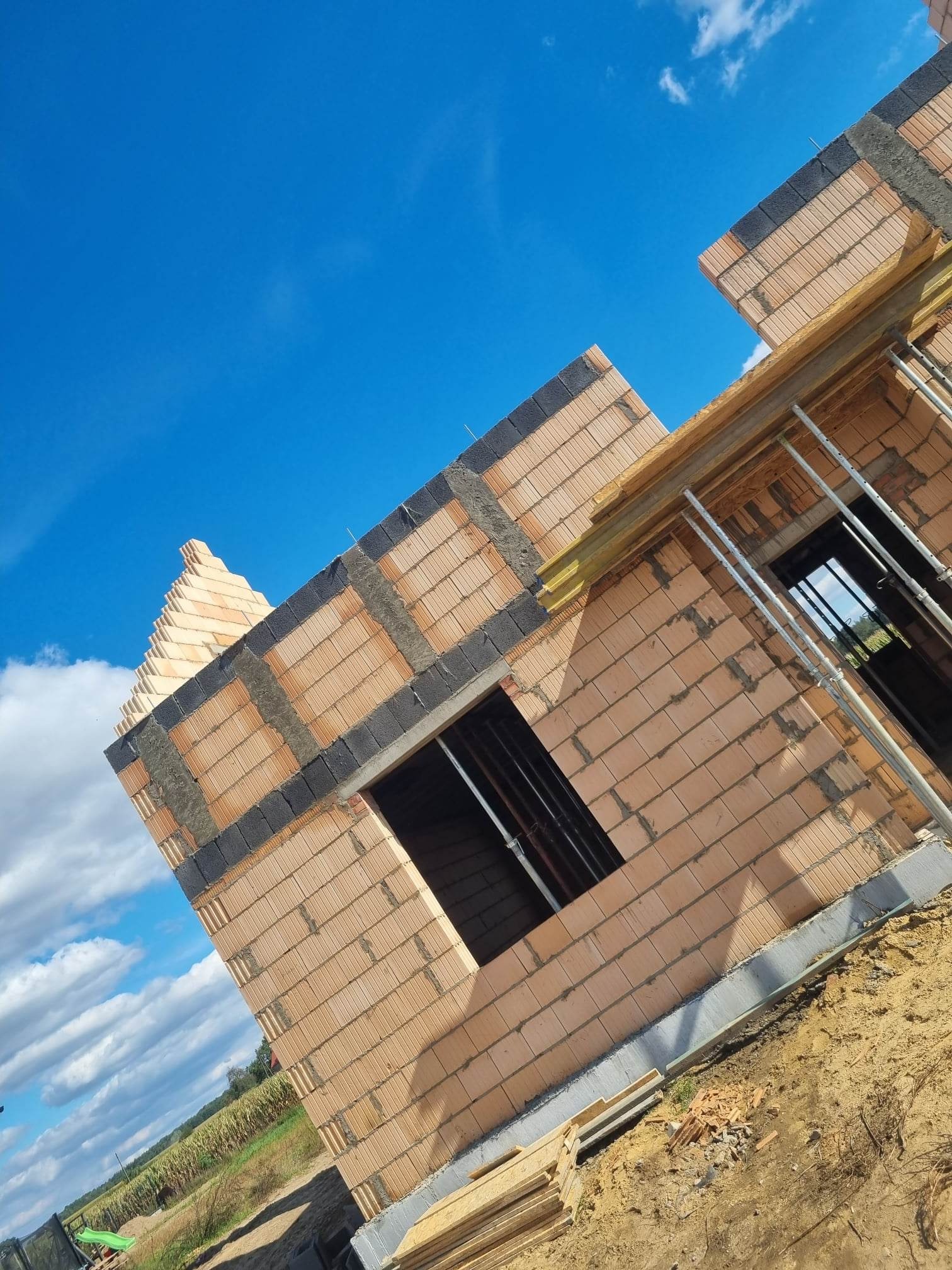 Wznoszenie ścian z cegły ceramicznej z widocznymi otworami okiennymi i betonowymi nadprożami na budowie w Nowym Tomyślu, na tle błękitnego nieba.