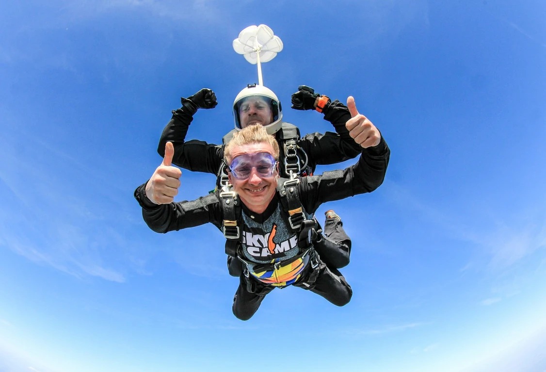 Uśmiechnięty mężczyzna w goglach i czarnym stroju do skoków spadochronowych, dający kciuk w górę podczas skoku w tandemie na tle błękitnego nieba.