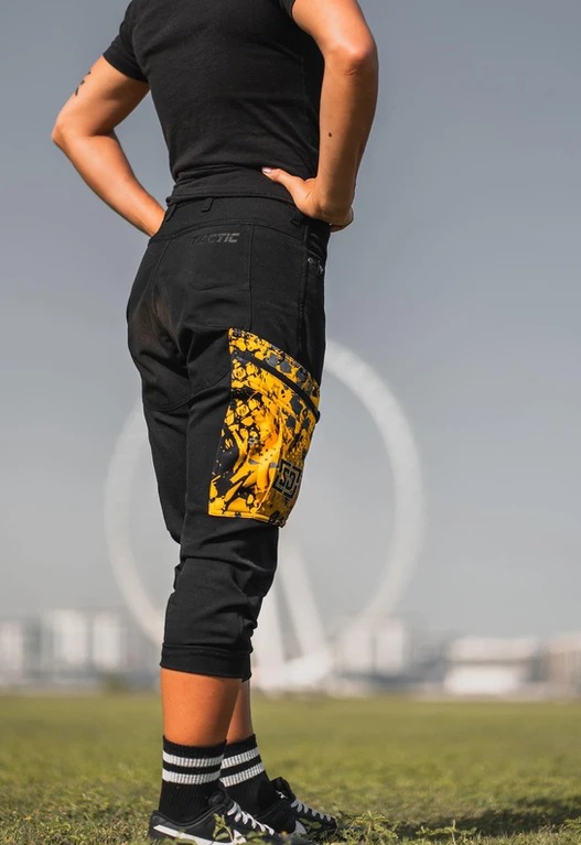 Czarna odzież robocza typu rybaczki z żółto-czarną kieszenią cargo na udzie, widoczne logo 'TACTIC' na tylnej kieszeni, na tle panoramy miasta z kołem obserwacyjnym.