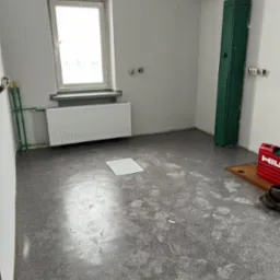 Pusty pokój w trakcie remontu z szarą podłogą, białymi ścianami, grzejnikiem, zieloną szafką i śladami po pracach budowlanych na podłodze.