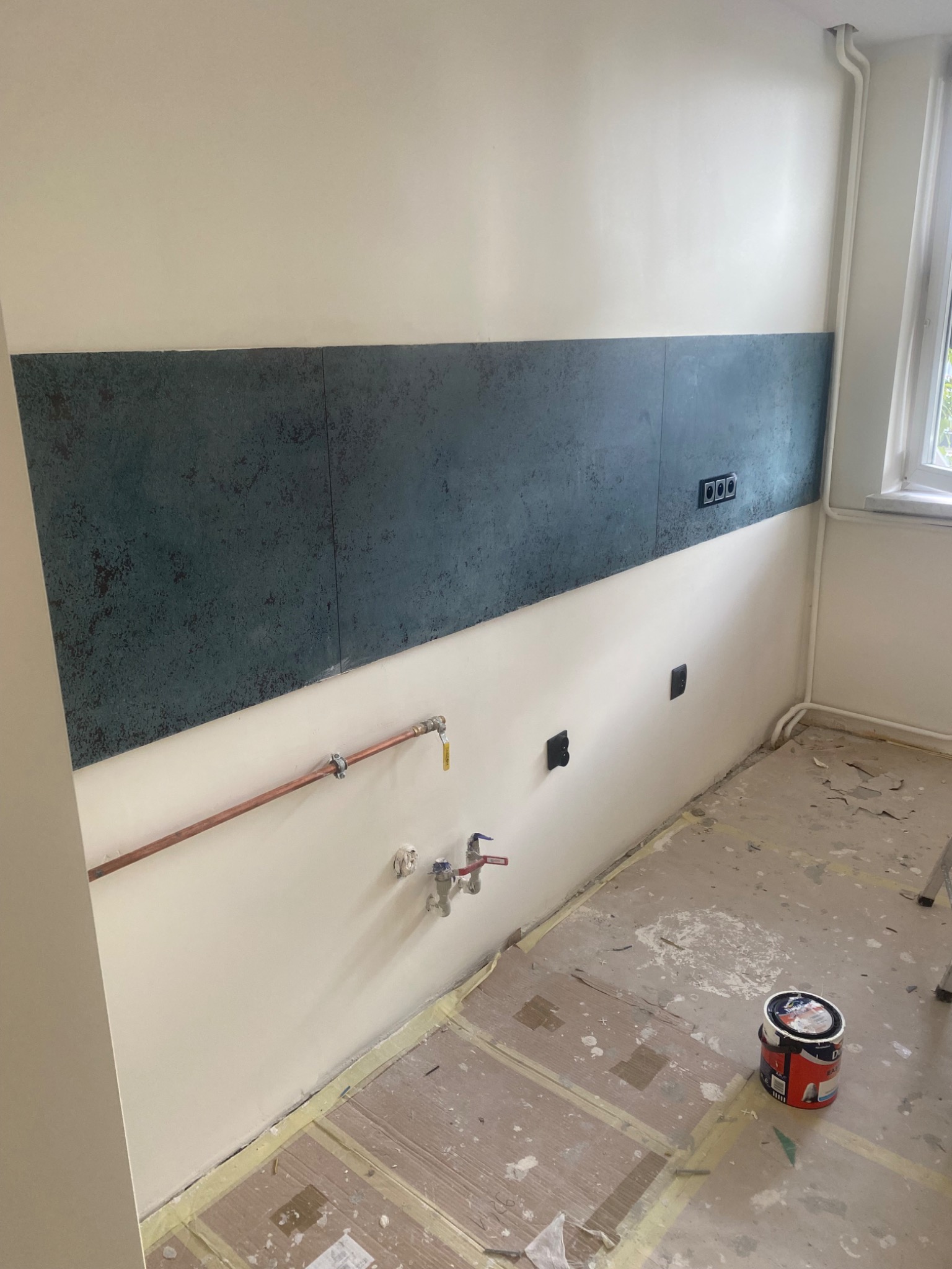 Fragment wnętrza w trakcie prac wykończeniowych: ściana z częściowo położoną dekoracyjną okładziną w odcieniach szarości i granatu, widoczne instalacje hydrauliczne z miedzianymi rurami, puszki...