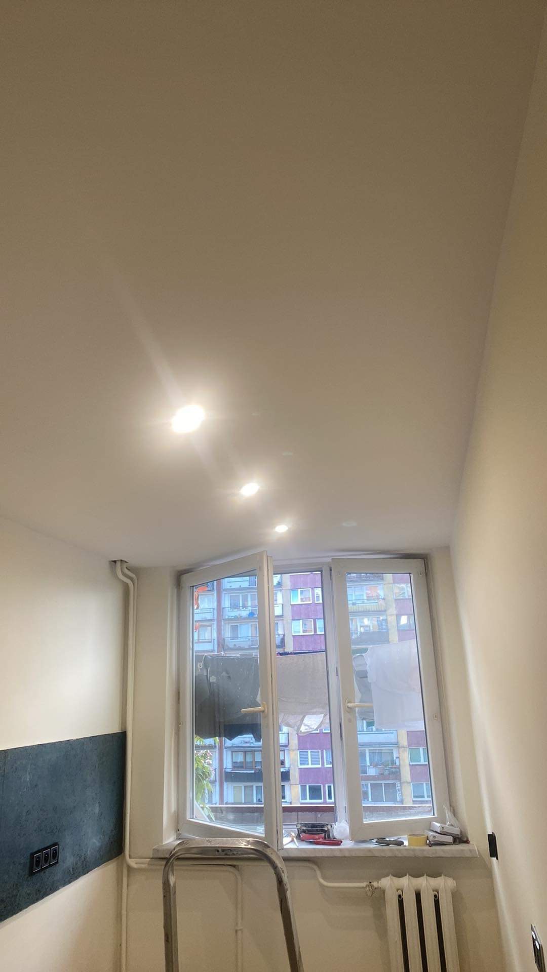 Biały sufit z zamontowanymi trzema okrągłymi lampami LED, otwarte okno z widokiem na blok mieszkalny, drabina oparta o ścianę, fragment ściany z dekoracyjną szarą okładziną oraz grzejnik pod oknem.