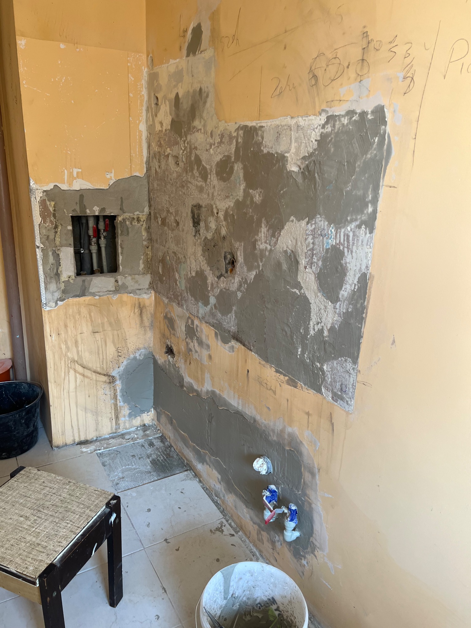 Ściana w trakcie przygotowywania pod wykończenie, widoczne fragmenty starej farby, nałożona warstwa szpachli, odsłonięte rury instalacji wodnej z zaworami, notatki wykonawcy na ścianie, wiadro...