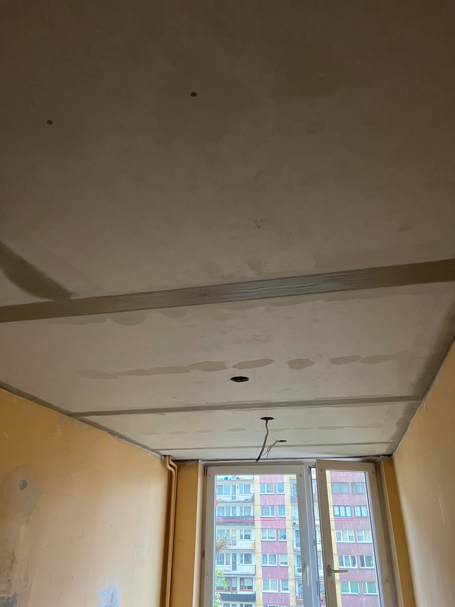 Pokój w trakcie prac wykończeniowych: surowy, szpachlowany sufit z widocznymi otworami na oświetlenie i wystającymi kablami, żółte ściany z miejscowymi łatami szpachli, okno z widokiem na blok...