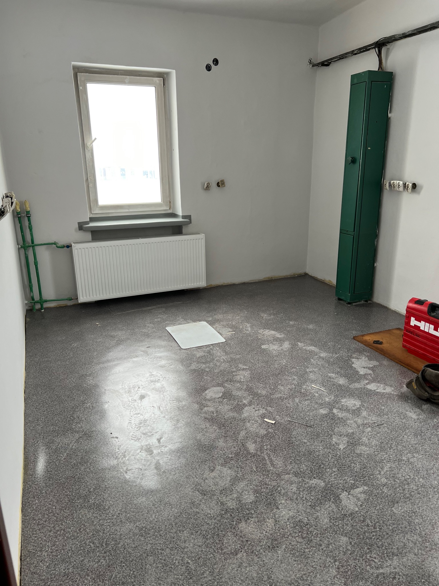 Pusty pokój w trakcie remontu z szarą podłogą, białymi ścianami, grzejnikiem, zieloną szafką i śladami po pracach budowlanych na podłodze.