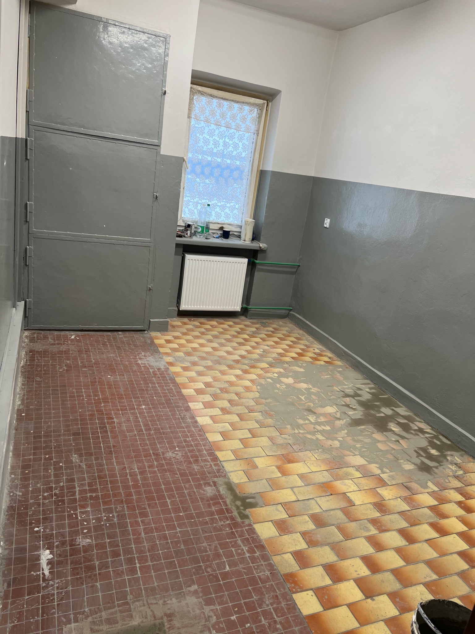 Pokój w trakcie renowacji z częściowo położonymi brązowymi i pomarańczowymi płytkami na podłodze, szarą szafką w rogu i grzejnikiem pod oknem.