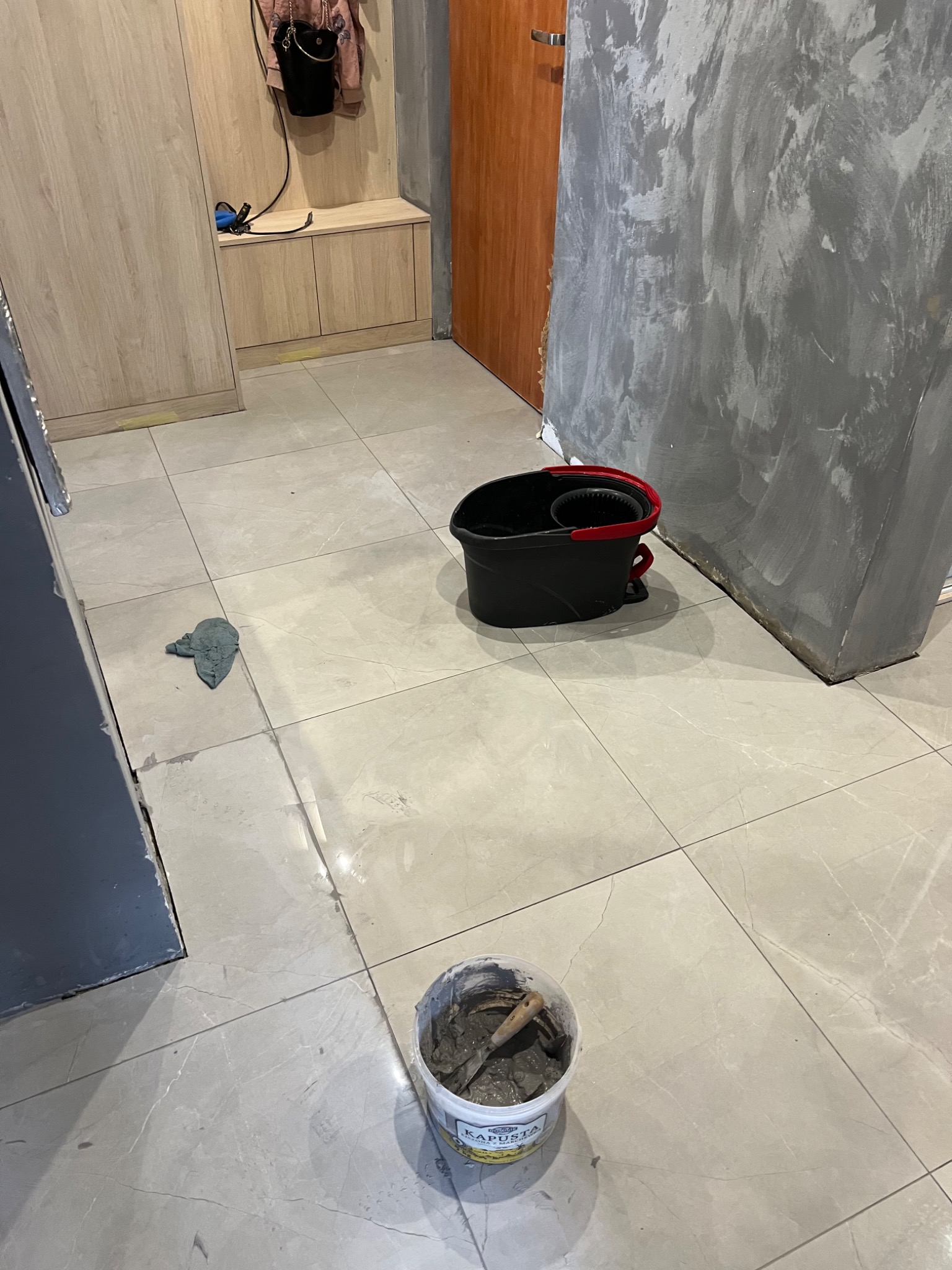 Szare płytki podłogowe w trakcie prac wykończeniowych, widoczne wiadro z zaprawą, czarne wiadro z wyciskarką do mopa i szmatka, ściana częściowo wykończona szarą farbą dekoracyjną.