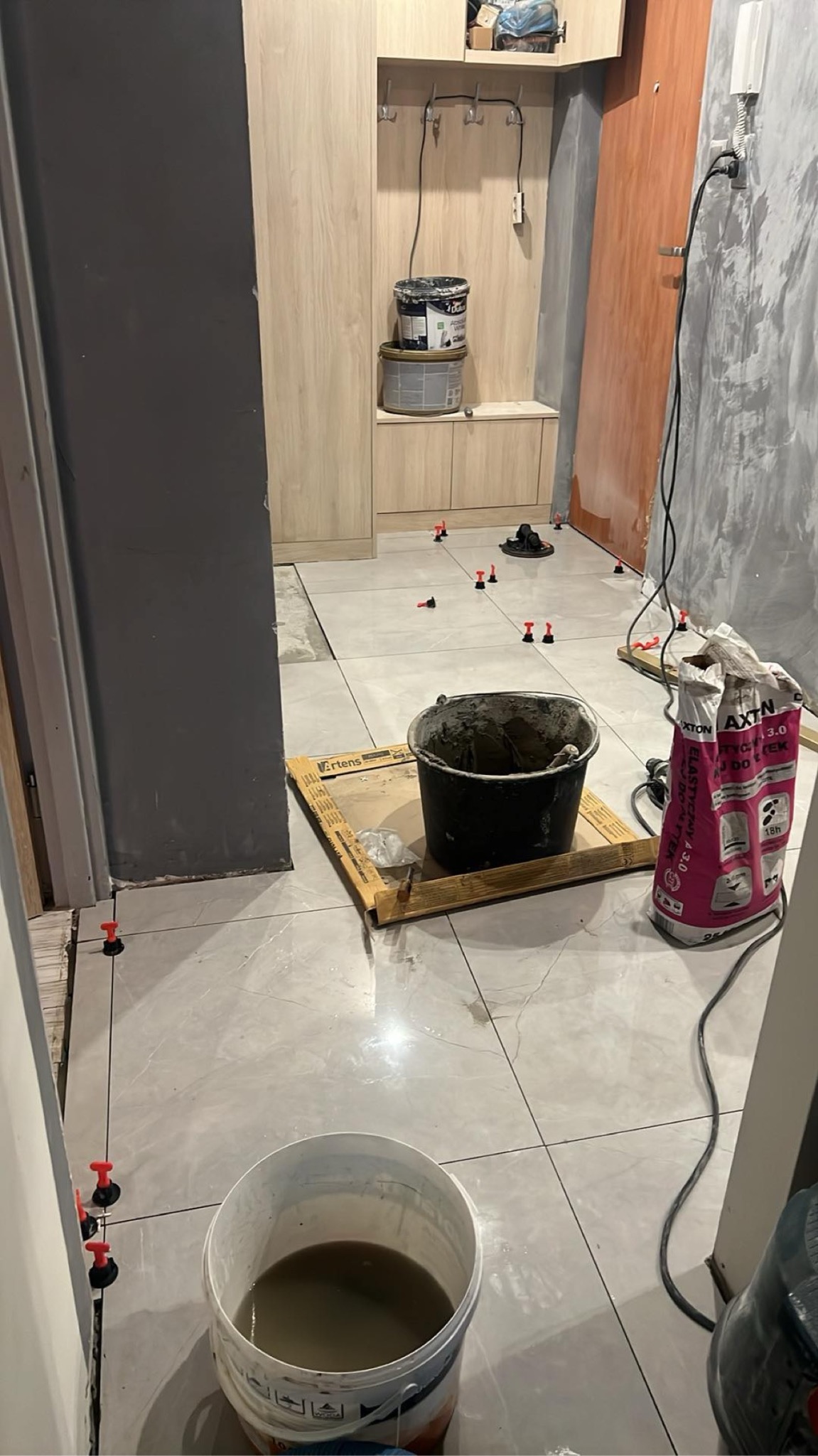 Układanie płytek podłogowych w przedpokoju, widoczne wiadra z zaprawą, paczka kleju Axton, system poziomowania płytek, szare ściany i jasne meble w tle.