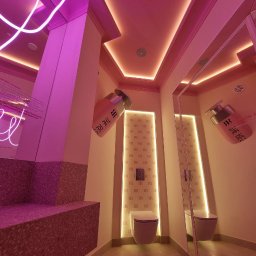 Vasyl Smachynskyi - Nietypowa łazienka w różowych odcieniach z neonowym oświetleniem, wzorzystą ścianą i dużymi flakonami perfum jako elementami dekoracyjnymi.