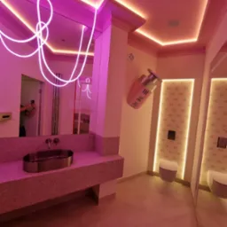 Awangardowa łazienka w różu: lustro z neonowym akcentem, umywalka z terazzo, toaleta i dekoracja ścienna w kształcie flakonu perfum. Nietypowy design.