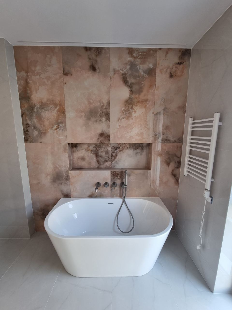 Nowoczesna łazienka z wolnostojącą białą wanną, mozaikową ścianą w odcieniach beżu i brązu oraz grzejnikiem drabinkowym na szarej ścianie.