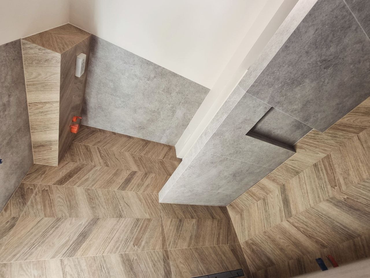 Ukończona łazienka z szarymi płytkami ściennymi i podłogą w jodełkę z płytek imitujących drewno, widoczna instalacja elektryczna i fragment odpływu liniowego.