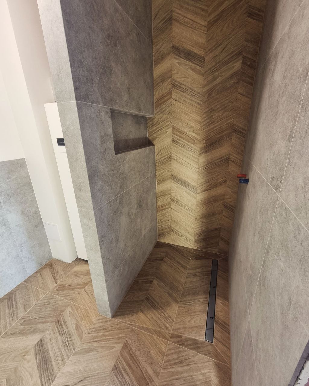 Ukończona łazienka z drewnopodobnymi płytkami ułożonymi w jodełkę na podłodze i ścianie, szara ściana z wnęką, odpływ liniowy w podłodze.