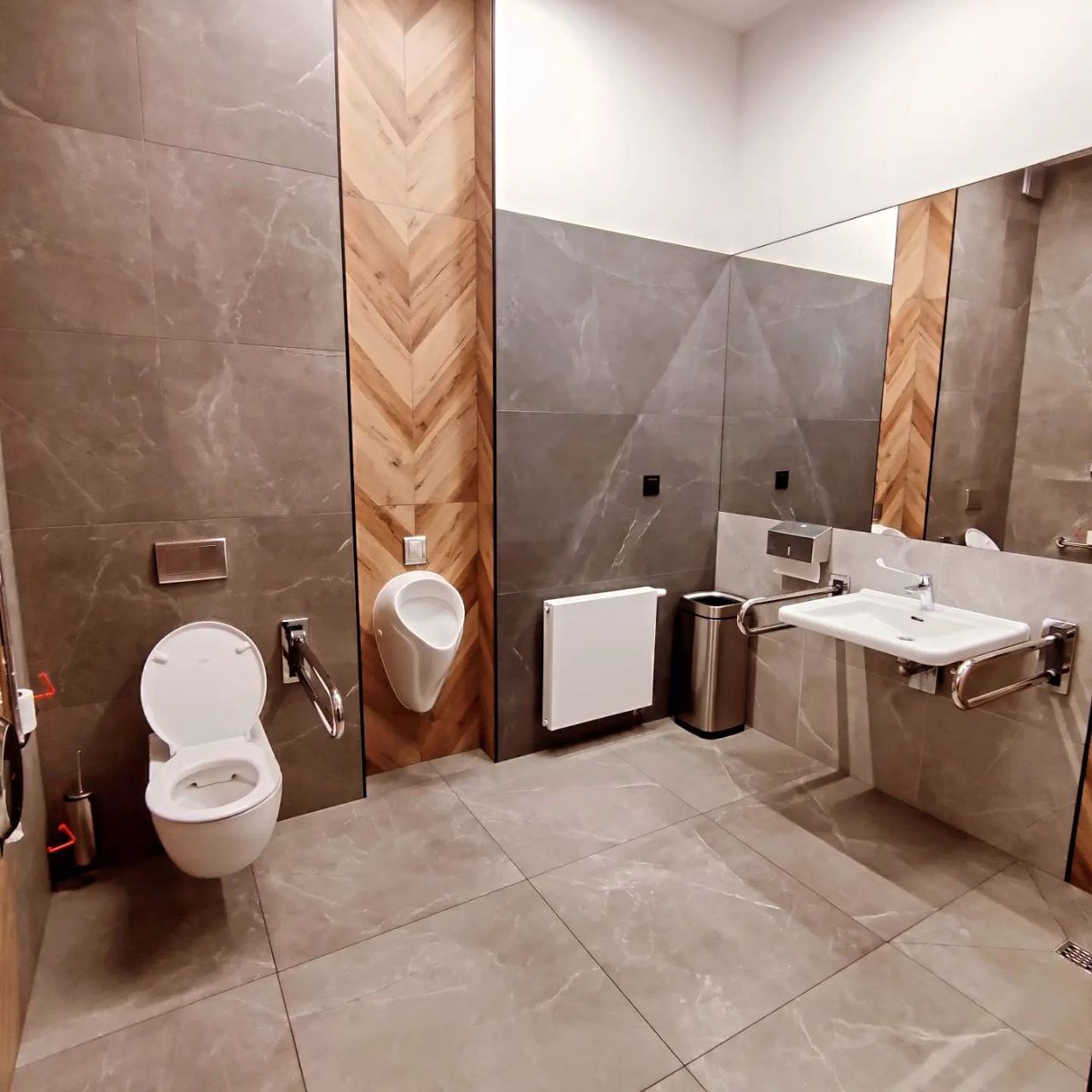 Nowoczesna toaleta publiczna z szarymi płytkami imitującymi marmur, drewnianą wstawką w jodełkę, wiszącą muszlą klozetową, pisuar i umywalką dostosowaną dla osób niepełnosprawnych, a także lustrem...