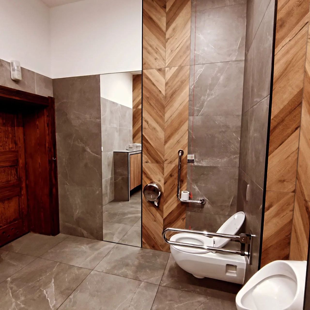 Wykończona łazienka z toaletą dla niepełnosprawnych, drewnopodobne panele ułożone w jodełkę na ścianie, szare płytki imitujące marmur, lustro wkomponowane w ścianę.