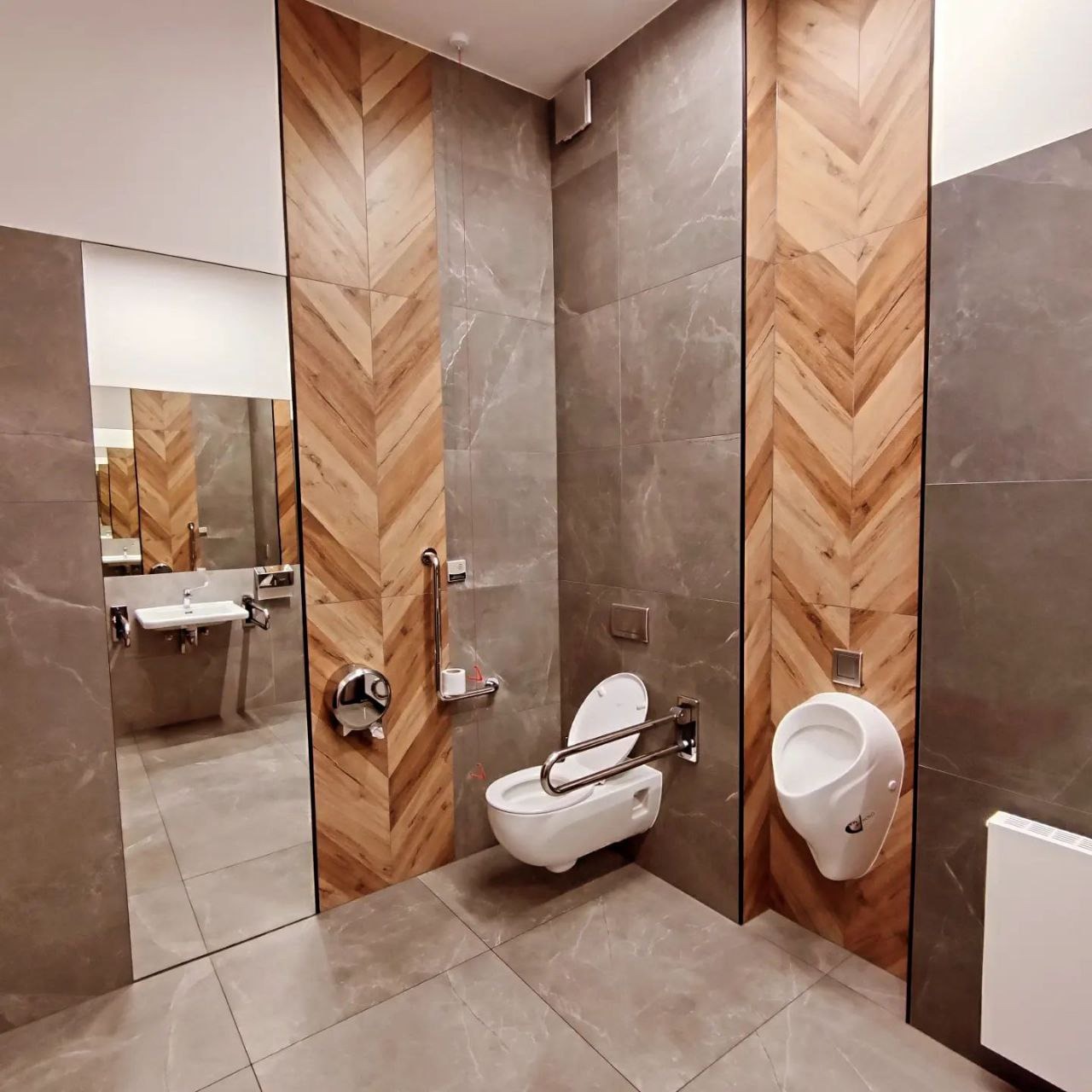 Nowoczesna toaleta publiczna z udogodnieniami dla niepełnosprawnych: wisząca miska WC z poręczami, pisuar, umywalka z lustrem, szare płytki imitujące marmur i drewniane panele w jodełkę.