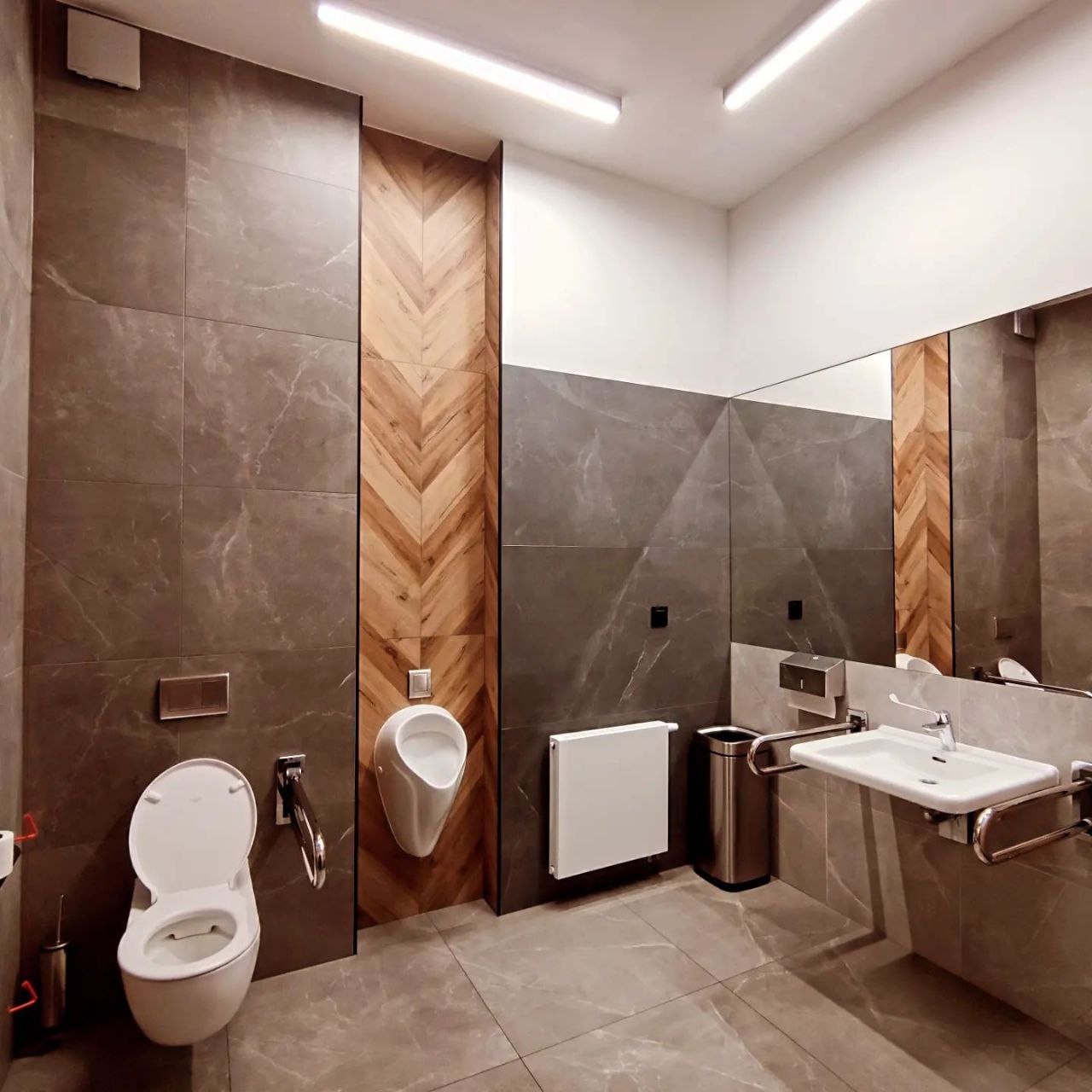 Publiczna toaleta z szarymi płytkami imitującymi marmur, drewnianą wstawką w jodełkę, umywalką dla niepełnosprawnych, pisuarami i toaletą.