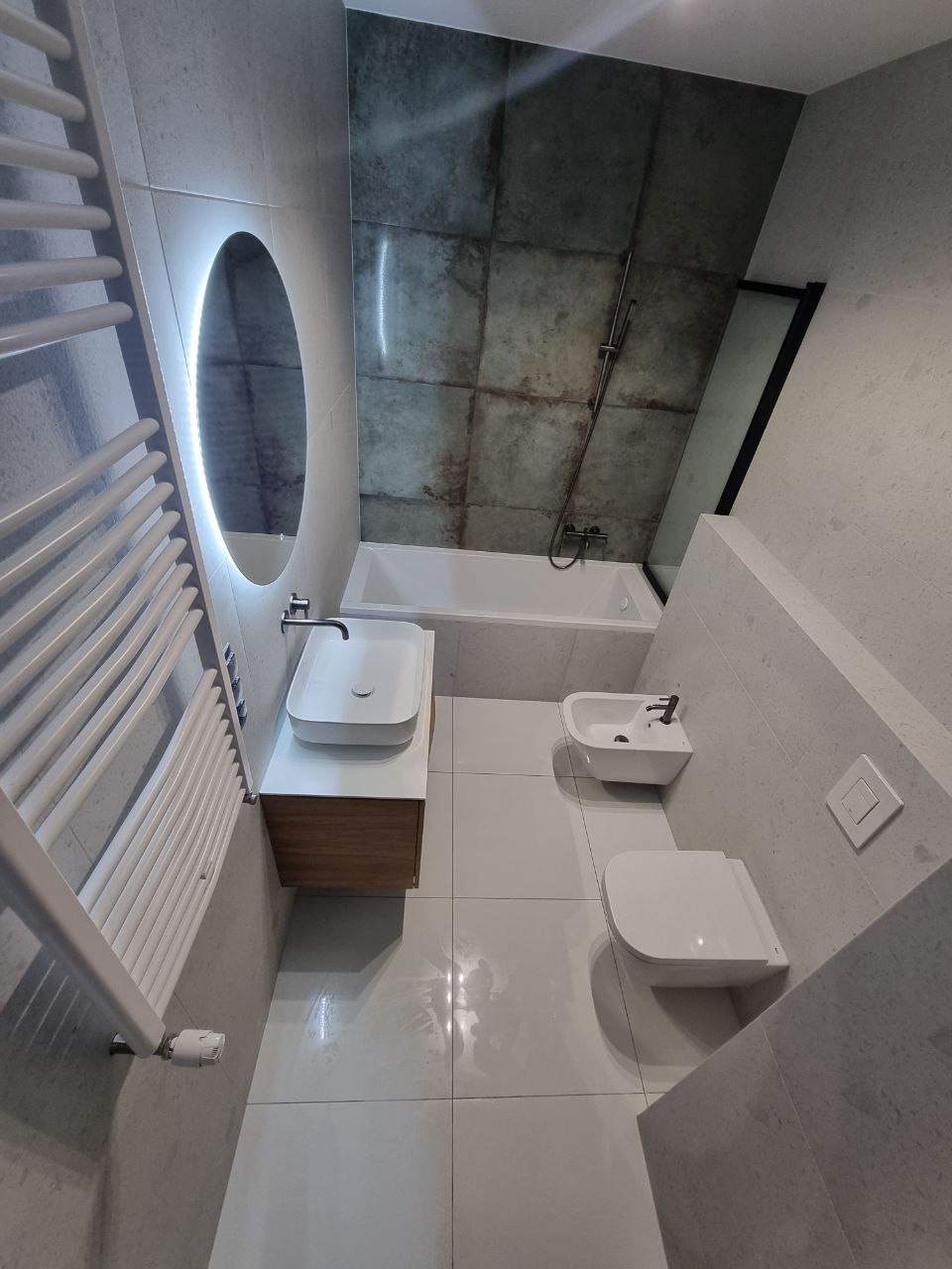 Nowoczesna łazienka z owalnym lustrem podświetlanym LED, białą umywalką nablatową z szafką z forniru, białą armaturą i płytkami imitującymi beton.