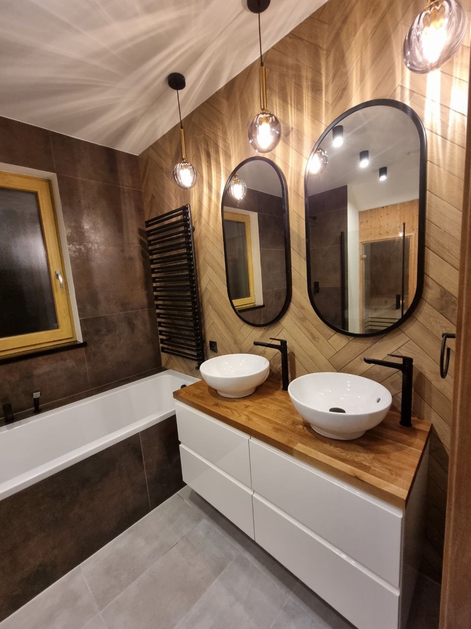 Nowoczesna łazienka z dwoma owalnymi umywalkami nablatowymi na drewnianym blacie, białymi szafkami, podłużnymi lustrami w czarnych ramach, dekoracyjnymi lampami wiszącymi i grzejnikiem drabinkowym...