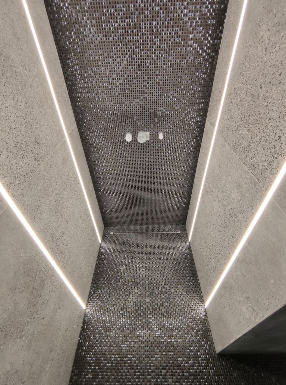 Wąskie wnętrze wykończone mozaiką w odcieniach szarości i oświetleniem LED, widok z dołu na sufit z trzema punktowymi lampami.