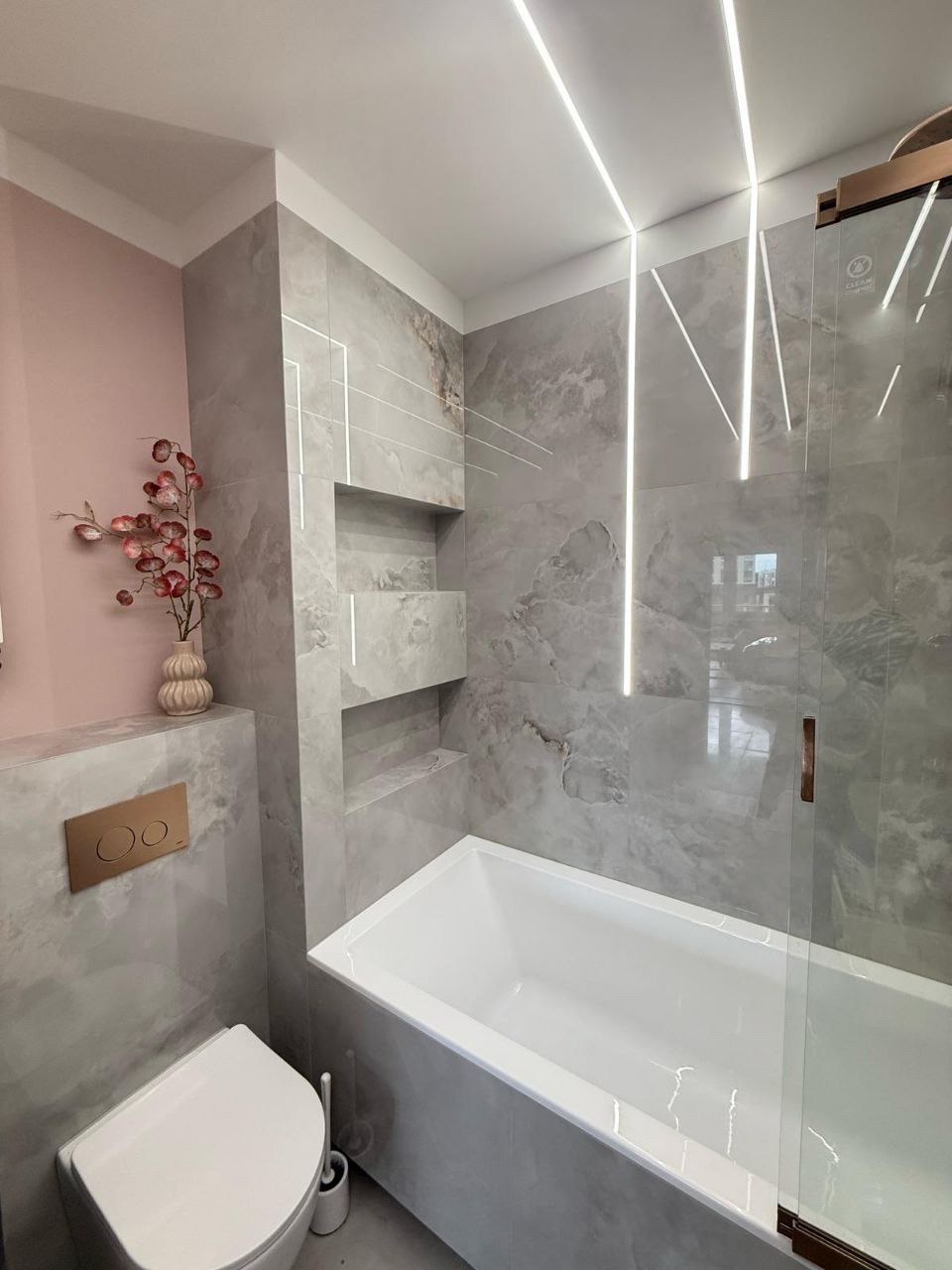 Elegancka łazienka w szarościach z akcentem pudrowego różu. Bateria w kolorze złota, podświetlane półki w ścianie, nowoczesna kabina i toaleta.