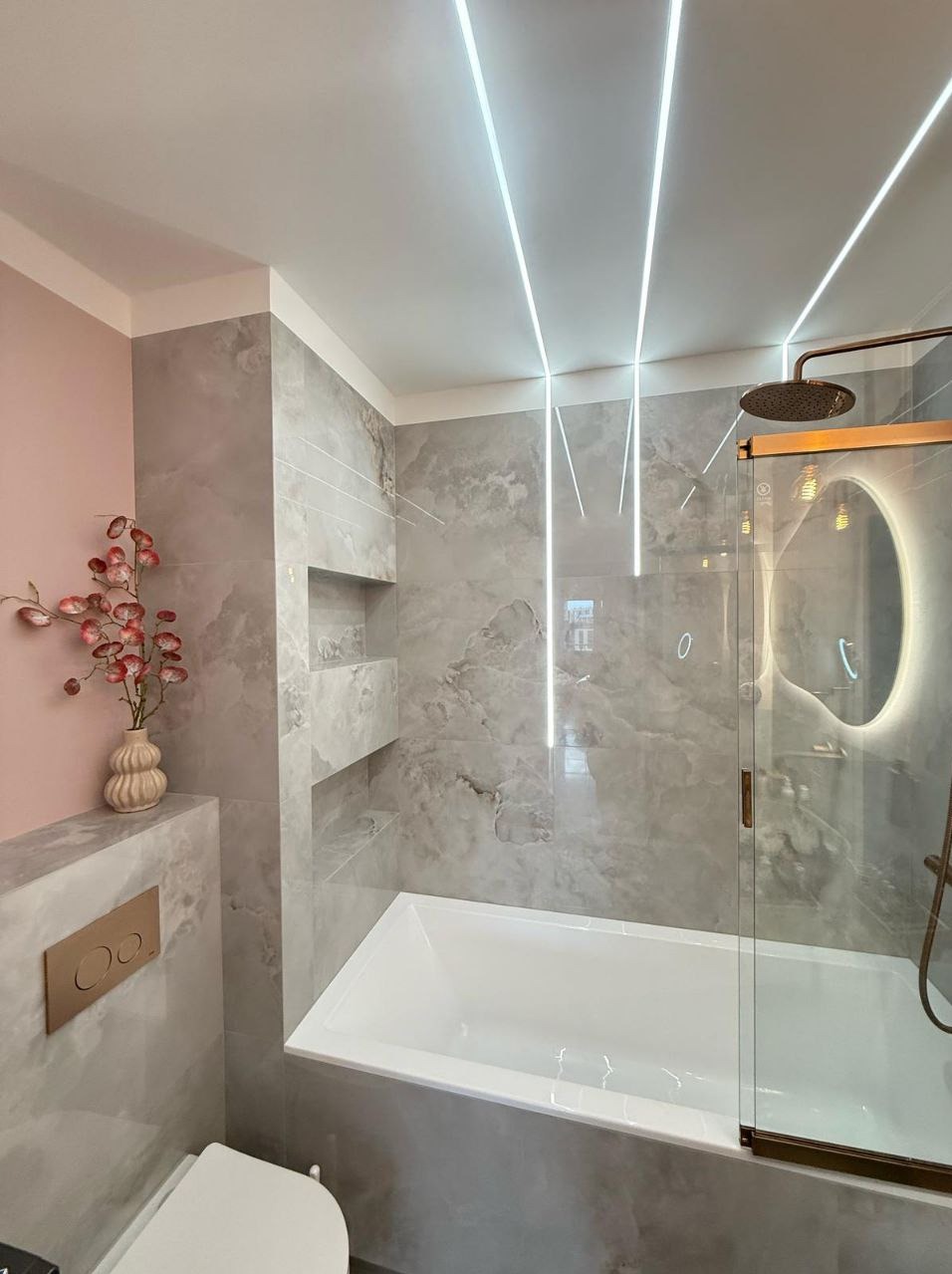 Nowoczesna łazienka z wanną, szarą marmurową okładziną i oświetleniem LED w suficie. Elementy dekoracyjne: wazon z kwiatami i okrągłe lustro.