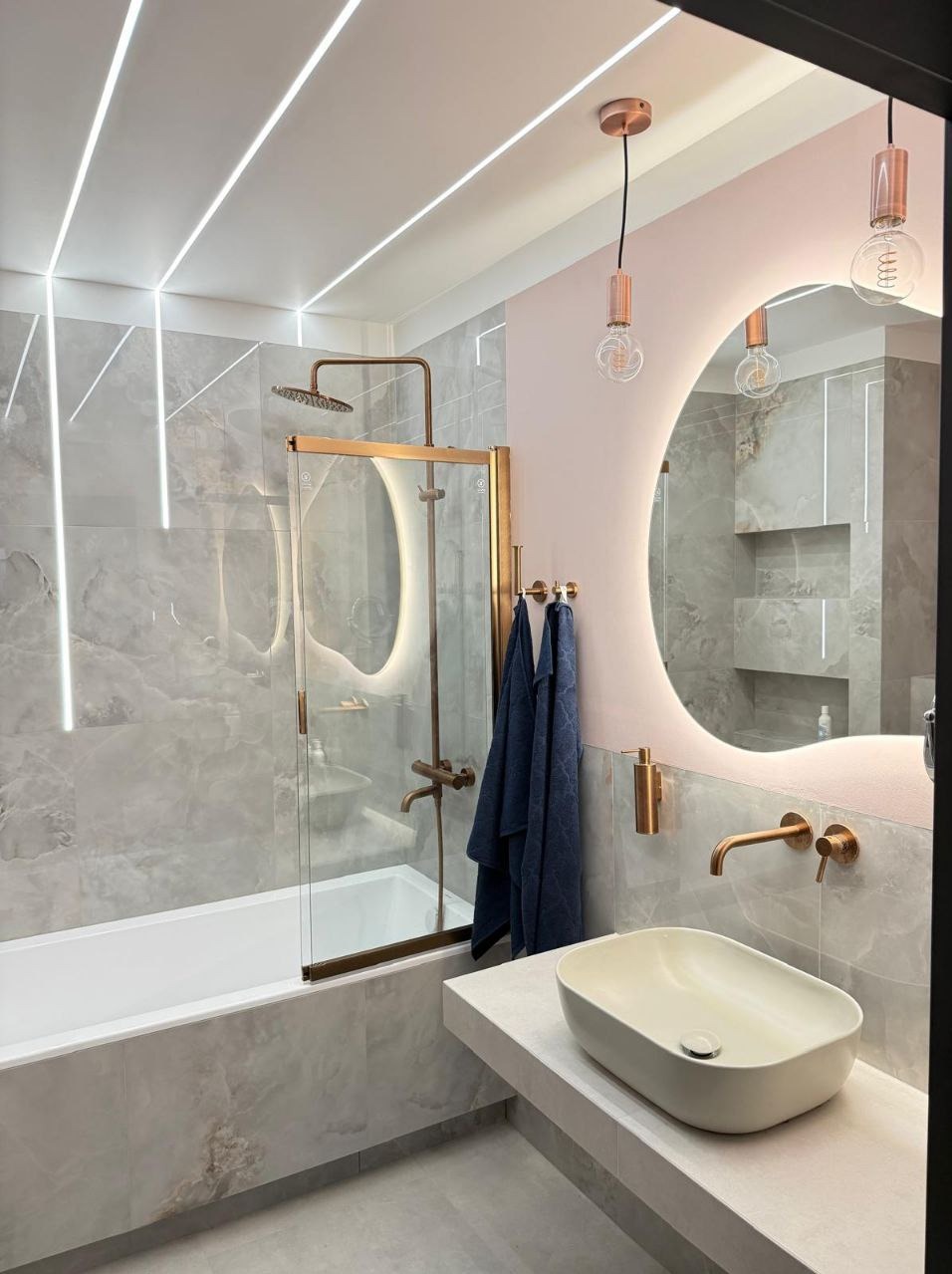 Nowoczesna łazienka z marmurowymi płytkami, podświetlanym lustrem i złotą armaturą. Minimalistyczny design z akcentem na detale i eleganckie wykończenie.