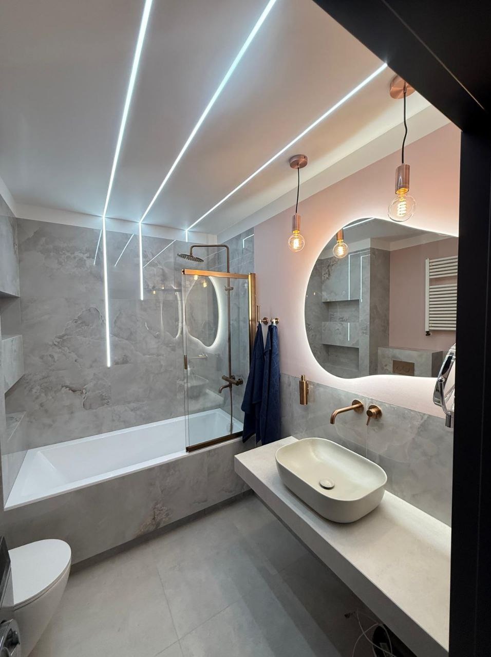 Elegancka łazienka z marmurowymi płytkami, podświetlanym lustrem i liniowym oświetleniem LED. Nowoczesna armatura w kolorze miedzi.