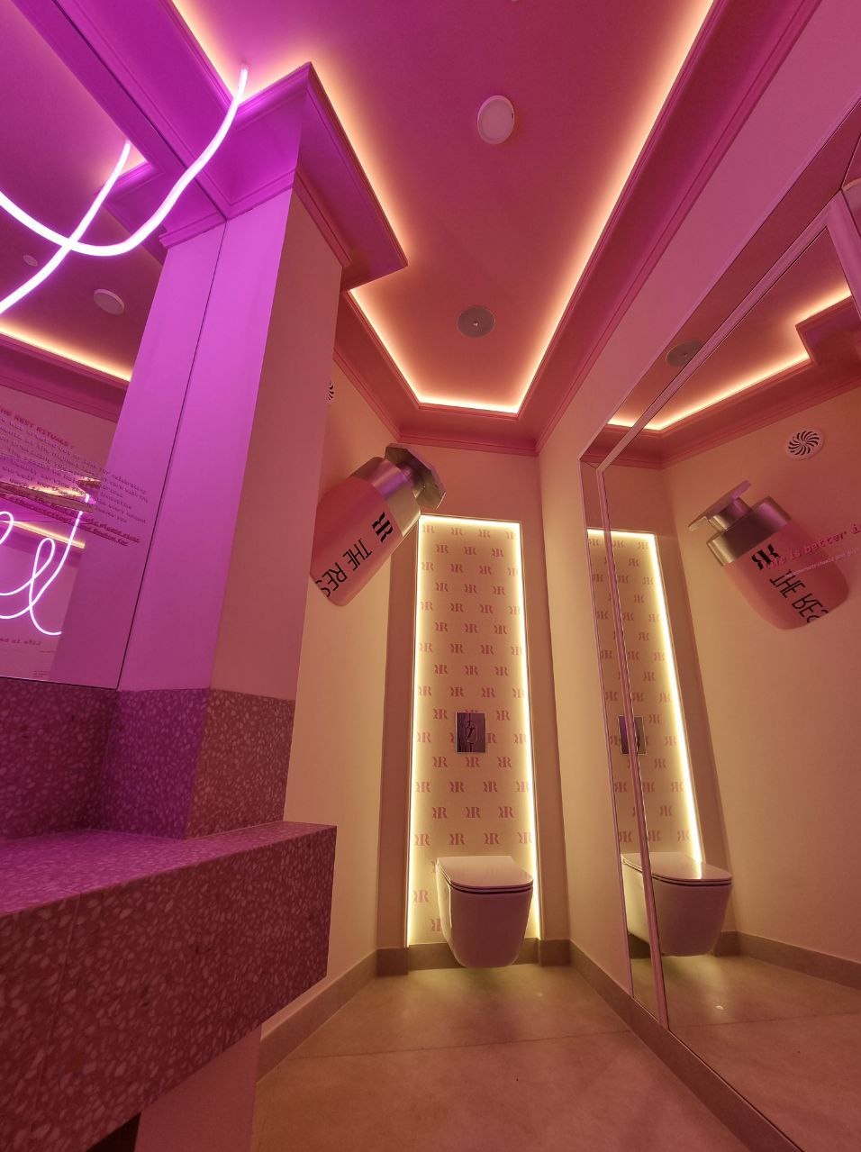 Nietypowa łazienka w różowych odcieniach z neonowym oświetleniem, wzorzystą ścianą i dużymi flakonami perfum jako elementami dekoracyjnymi.