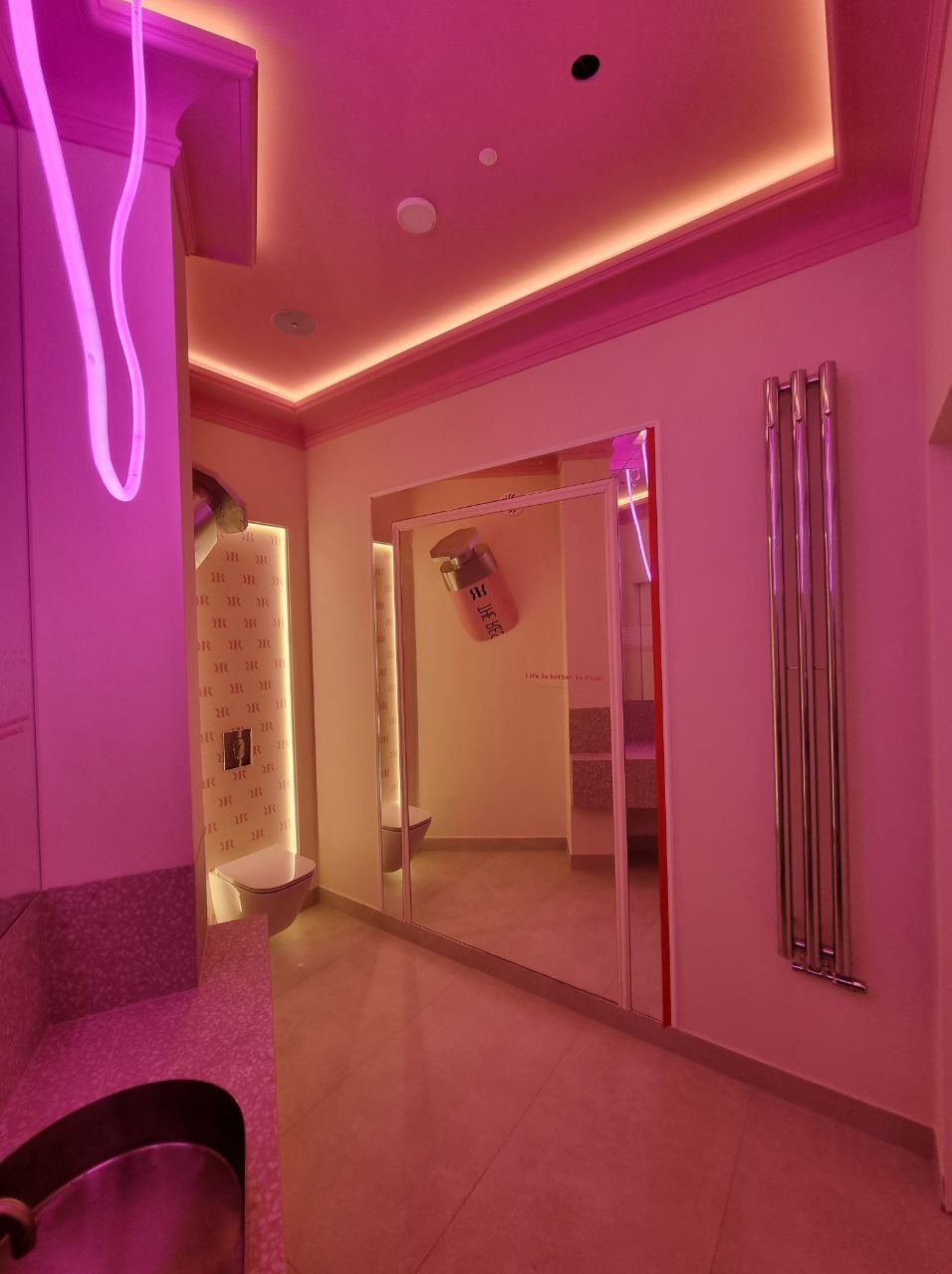 Awangardowa łazienka z neonowym oświetleniem i lustrami, odbijającymi wnętrze. Umywalka z wzorem, grzejnik drabinkowy, toaleta w tle. Całość w różowej tonacji.