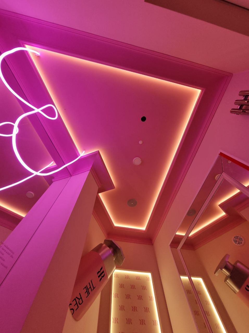 Nowoczesna łazienka z podświetlanym sufitem w odcieniach różu i beżu, dekoracyjnym neonem i dużymi imitacjami kosmetyków. Nietypowy design.