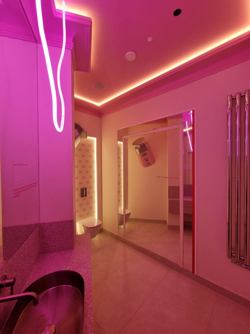 Nowoczesna łazienka w różowym oświetleniu neonowym. Lustrzane drzwi, dekoracyjna ściana z motywem 'R', designerska umywalka, grzejnik drabinkowy.