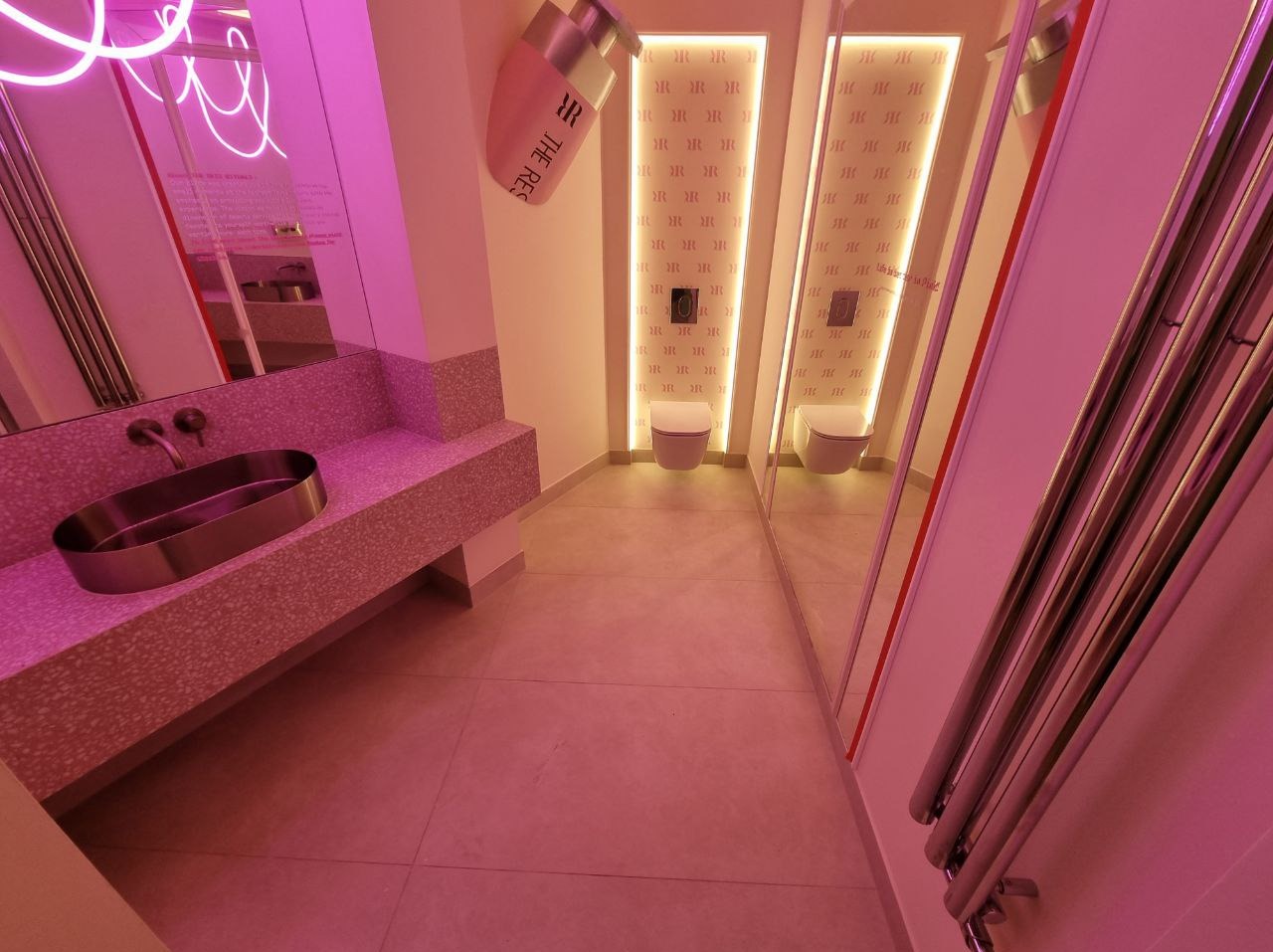 Nowoczesna toaleta z umywalką na blacie z lastryko, lustrami i neonowym oświetleniem w odcieniach różu. Widoczne detale wykończeniowe i armatura.