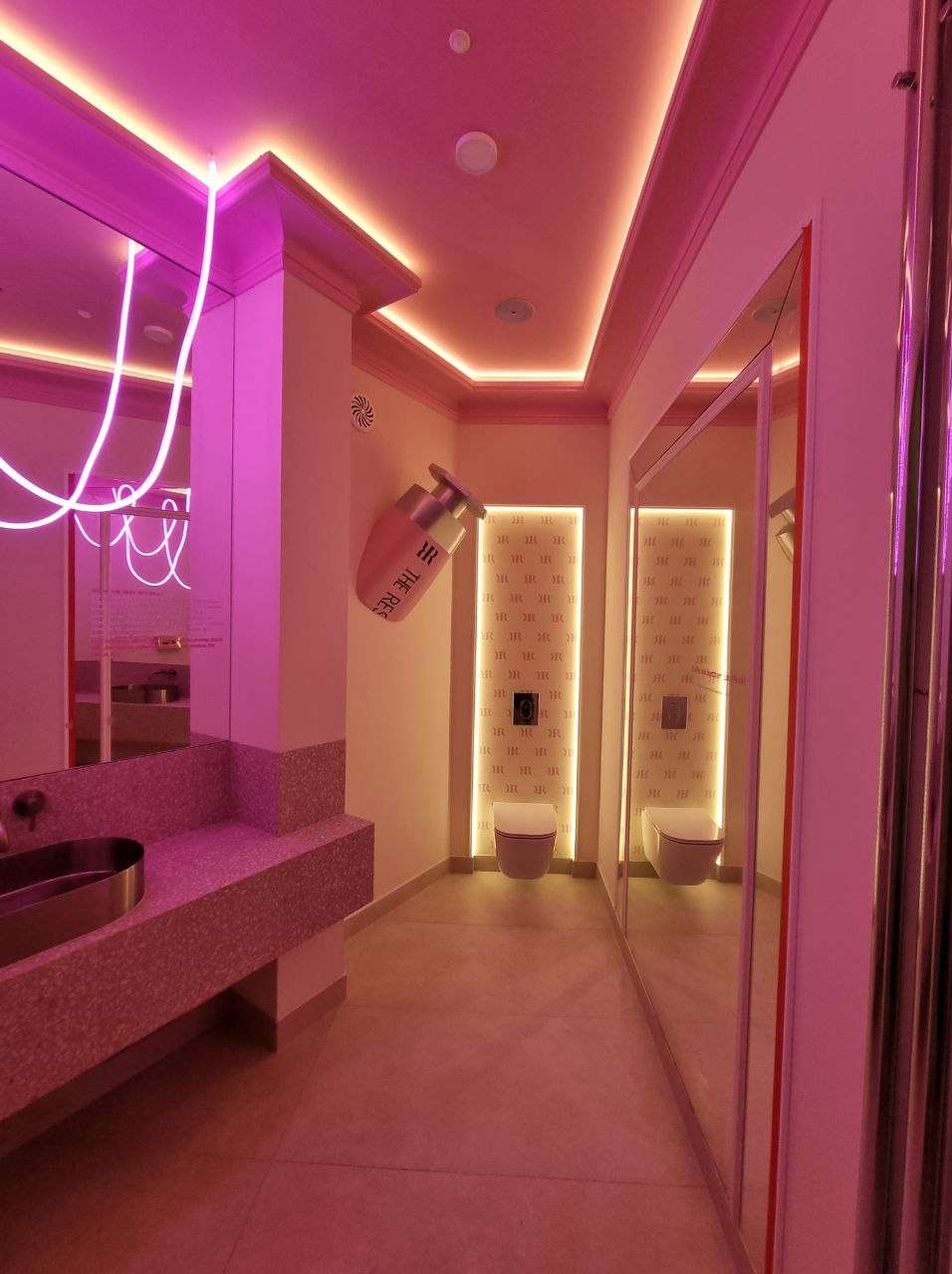 Różowa łazienka z neonami i designerską umywalką. Na ścianie dekoracja w kształcie butelki perfum. W tle toalety z podświetleniem i lustro.