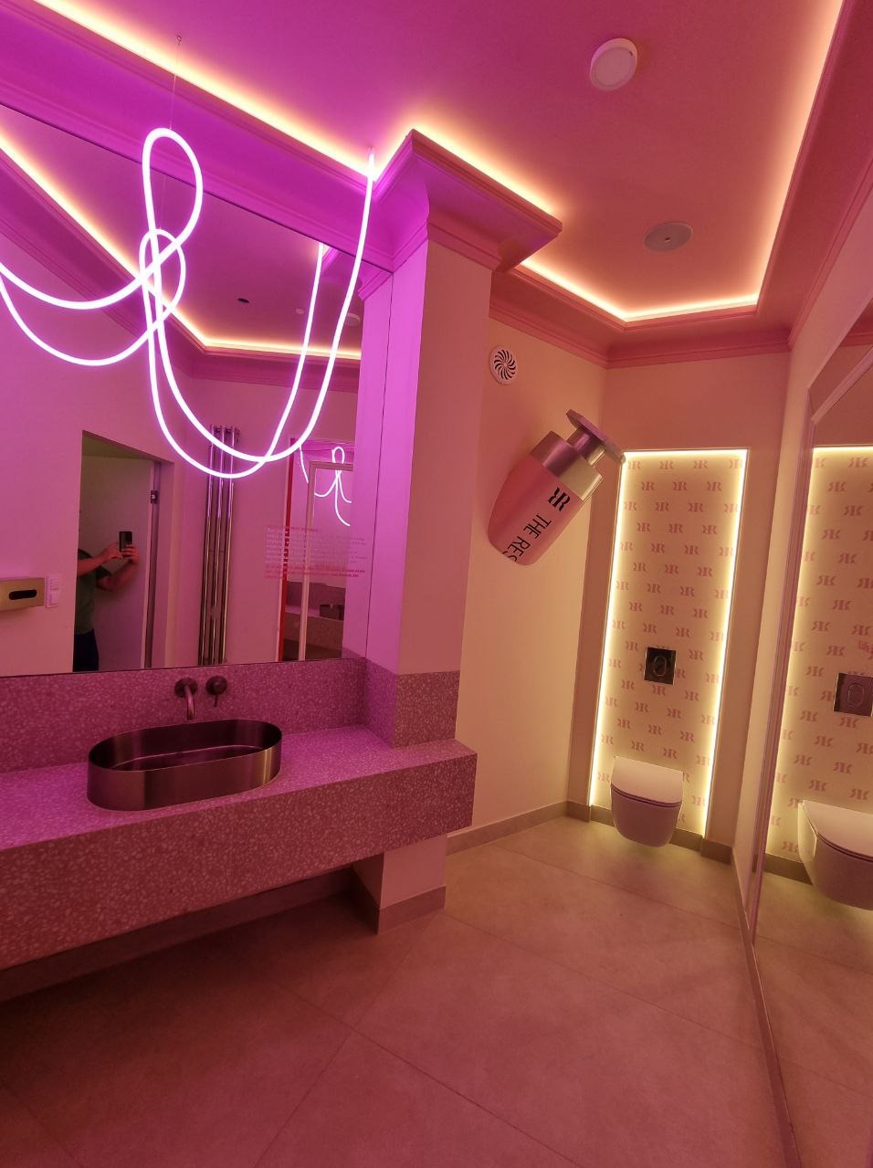 Awangardowa łazienka w różu: lustro z neonowym akcentem, umywalka z terazzo, toaleta i dekoracja ścienna w kształcie flakonu perfum. Nietypowy design.