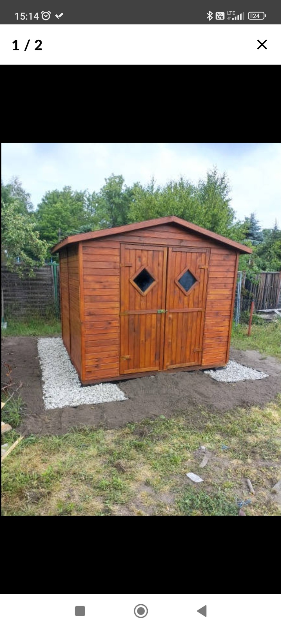 Drewniana altana ogrodowa z dwuspadowym dachem i podwójnymi drzwiami z romboidalnymi okienkami, otoczona fragmentami wysypanego żwiru.